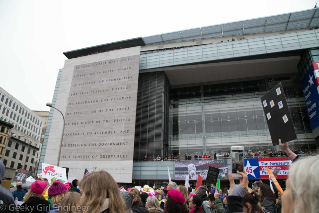 An interesting contrast of people exercising their First Amendment Rights, in front of the Newseum with the First Amendment written on the side of the building with people cheering from the building with a "Welcome President Trump" sign